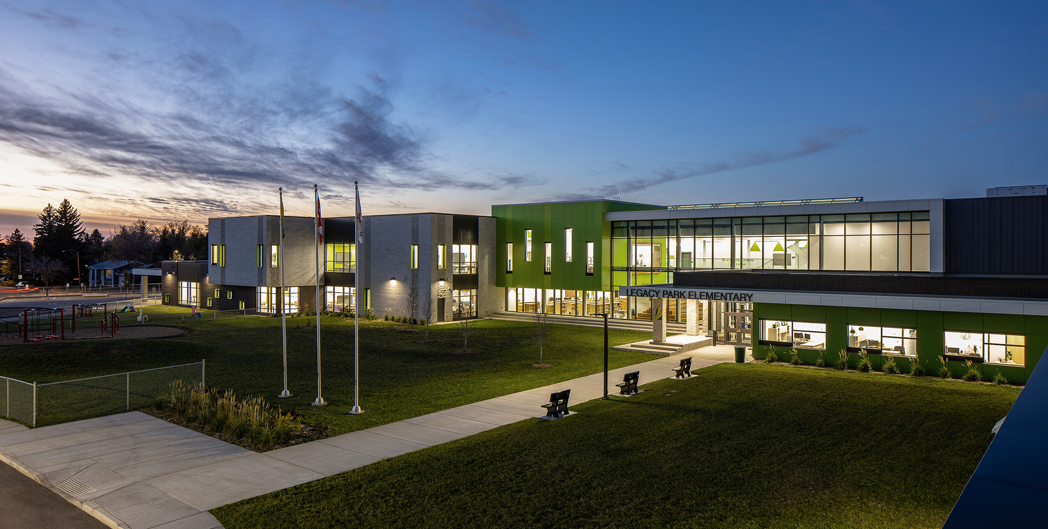 Exterior shot of the school in the evening time. You can see three flags in the front lawn and the lights are glowing through the windows of the building.
