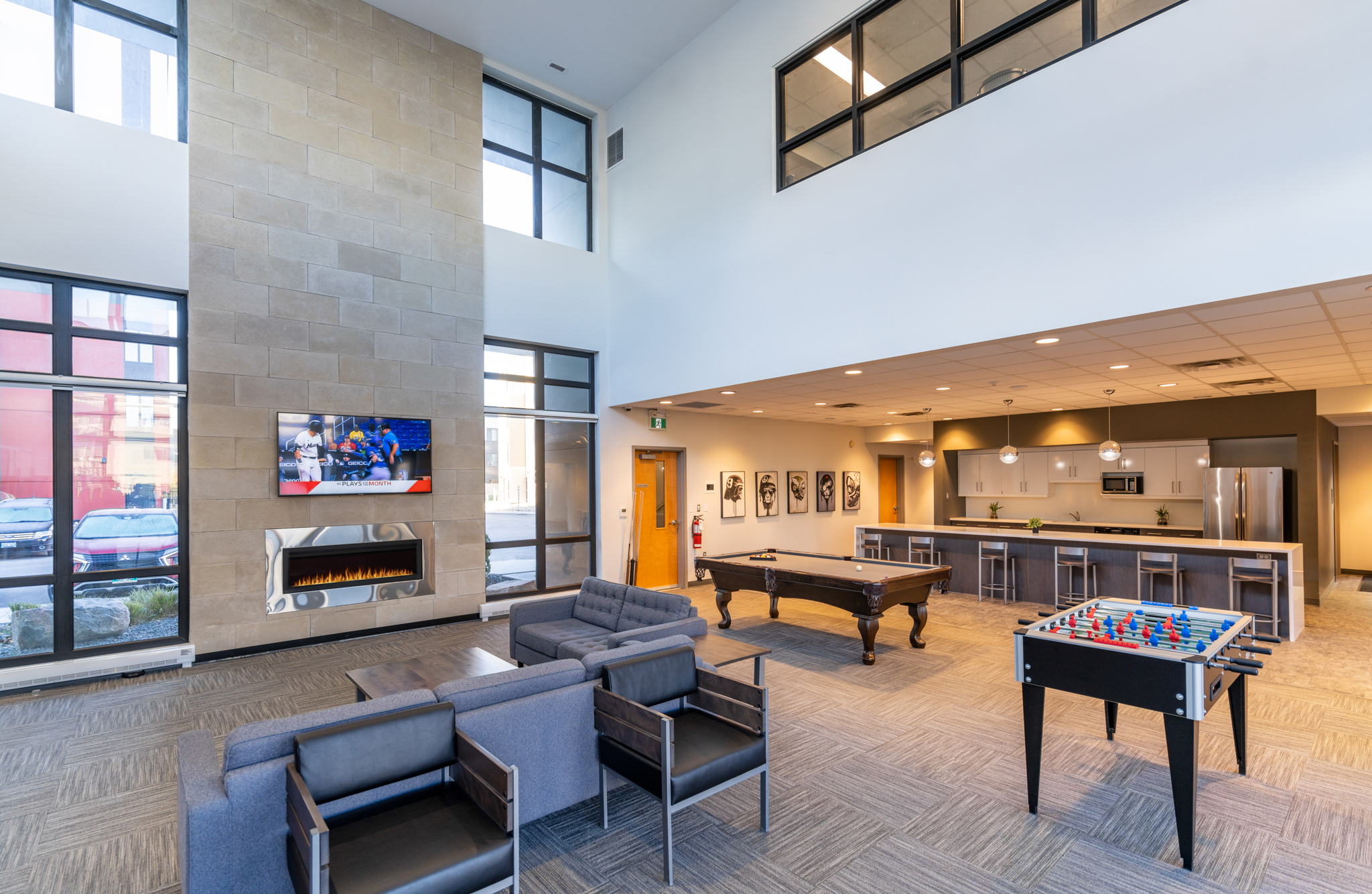 Image inside the lobby of the apartment block. There are couches, a large island with chairs, a fridge and a sink. There is a pool and foosball table, as well as an electronic fireplace and tv on the wall.