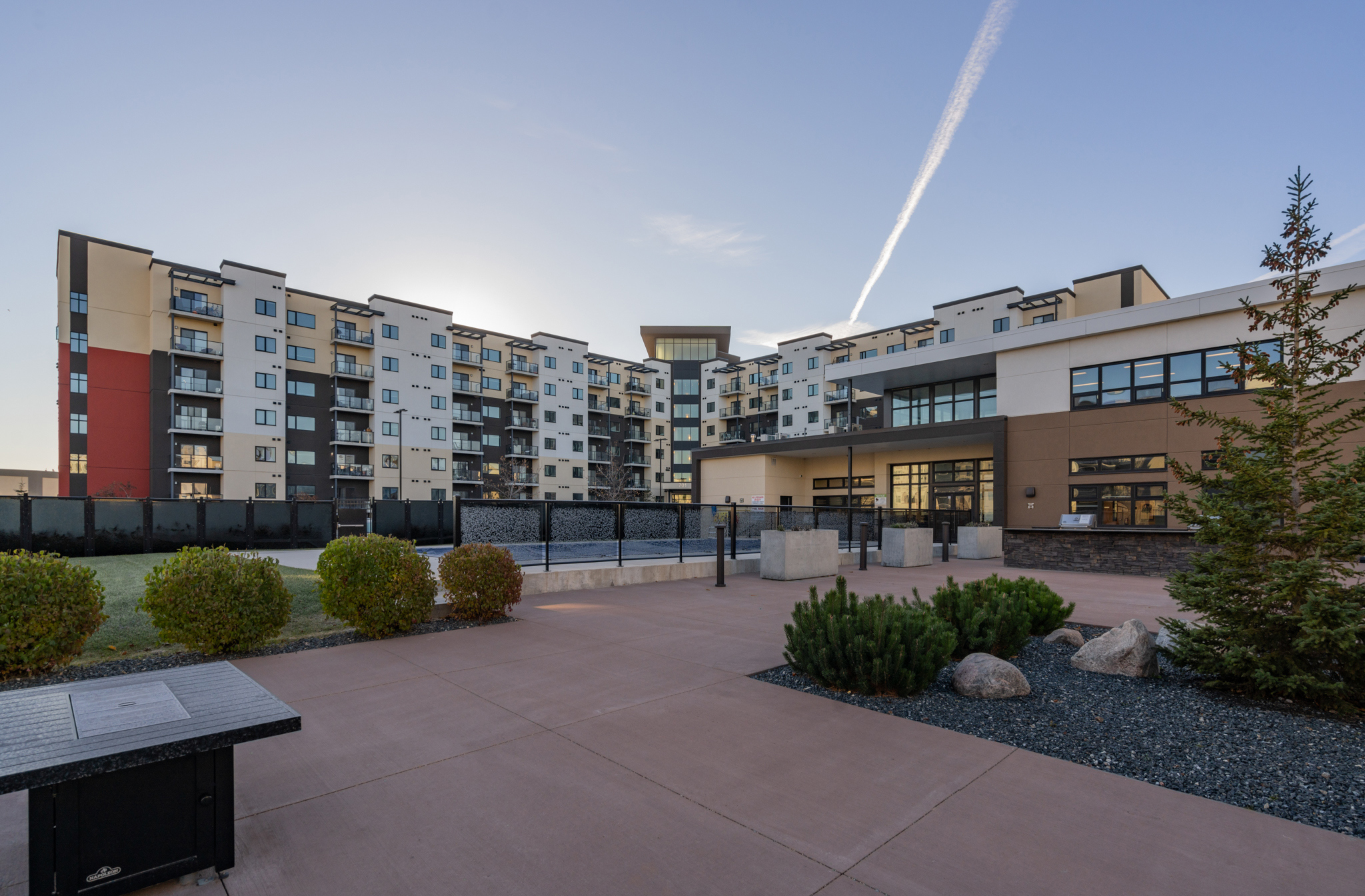 Image of the exterior of the apartment building. There is a courtyard with bushes and trees in front of the main entrance.