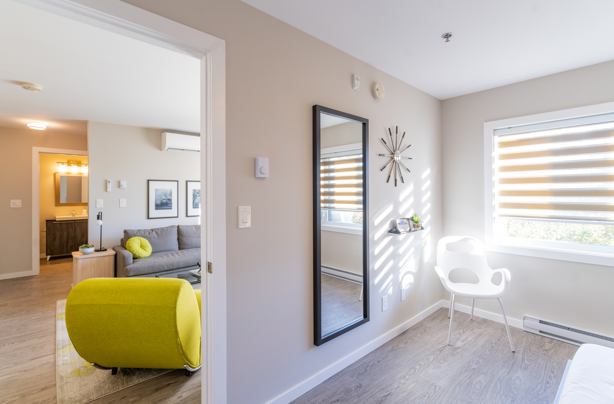 Image from inside of the bedroom of one of the suites. There is a long mirror on the wall and a white chair in the corner of the room. There is a starburst clock on the wall and a big window lets natural light spill into the room.