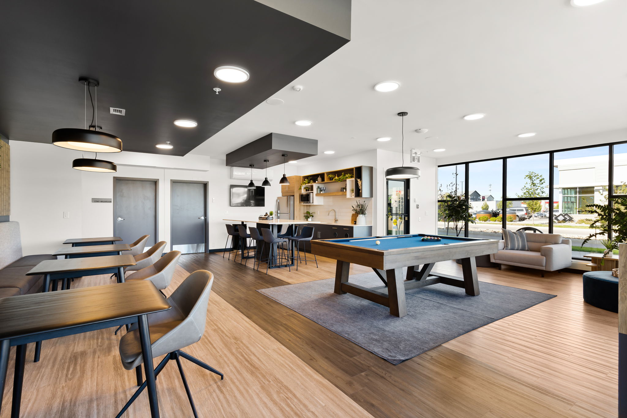 Image of a communal lounge on the ground floor. There is booth seating with four tables and chairs against it. There is a pool table, an island, a fridge, sink and other seating.